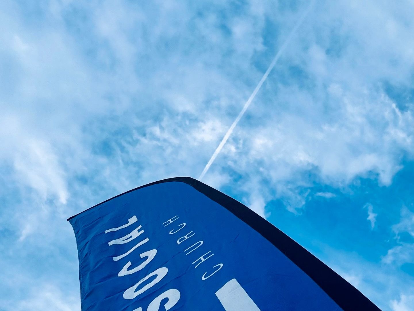 Blue Social Church flag against a bright sky with clouds and a contrail.